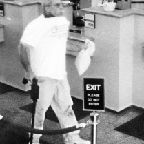 Brian Wells heads out of the PNC Bank in the Summit Towne Centre after robbing it of $8,702. Wells is carrying the cash in a white canvas bag; in his left hand is a homemade cane-shaped shotgun. He is sucking on a lollipop he grabbed while in the bank. The collar bomb is protruding from under his white T-shirt. He had demanded $250,000 from the chief teller, but left with whatever money she could give him. ERIE TIMES-NEWS, via FBI